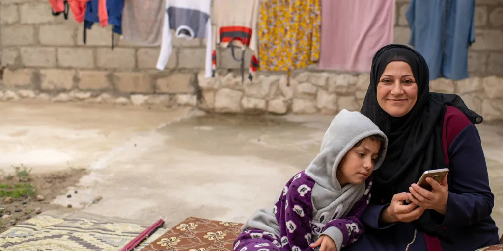 Nawal smiles at the camera while her daughter looks at a smart phone Nawal is holding.